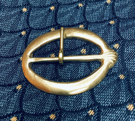 Brass girdle buckle with scallop pilgrim symbol and incised decoration, 5cm in length, displayed on dark fabric background.